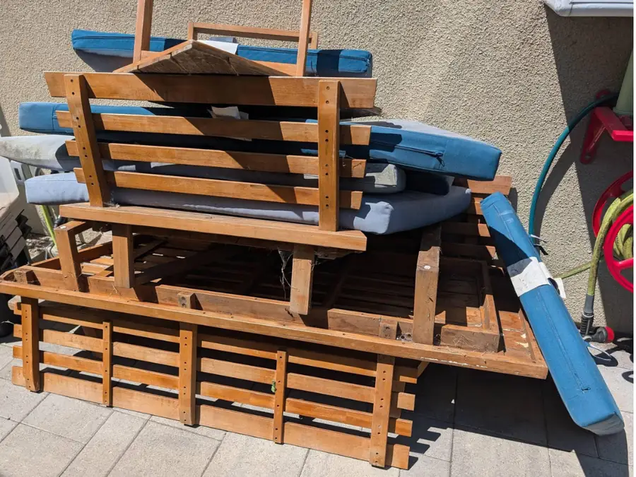 A stack of broken wooden patio furniture and cushions piled against a wall