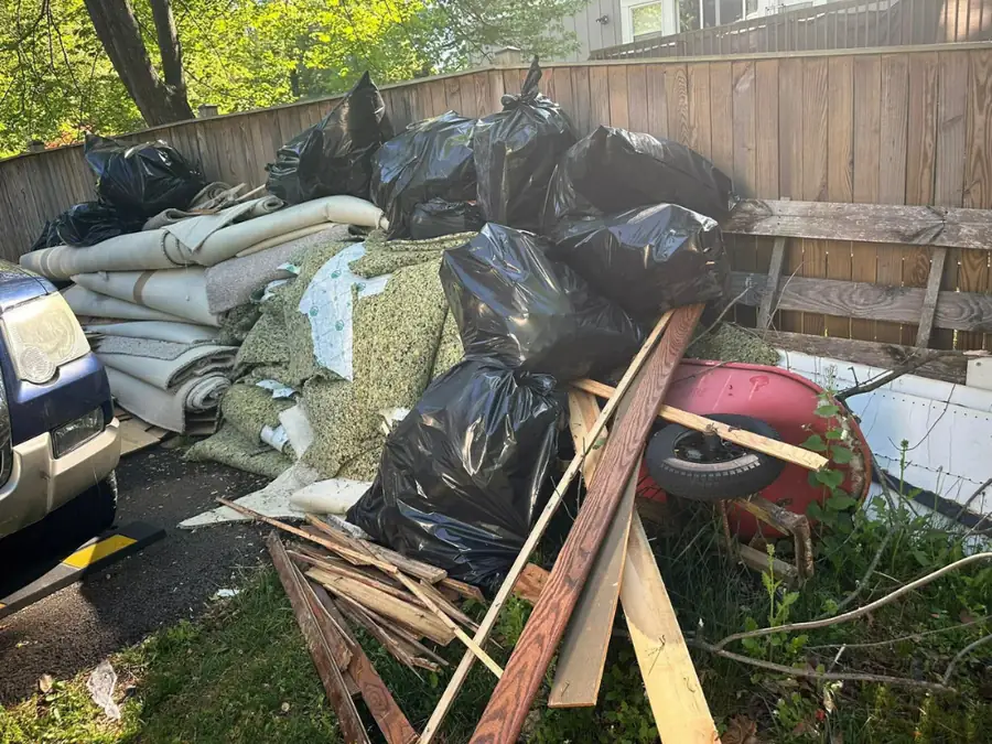 Rolled-up old carpet, underlay and bagged waste piled beside a fence